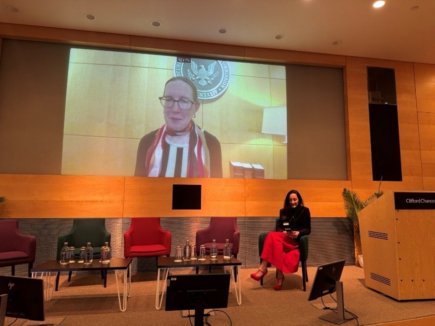 Hester Peirce on a screen in conversation with Emma Joyce in a conference room
